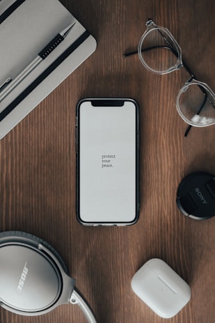 A neatly arranged flat lay on a wooden surface includes a smartphone displaying the message 'protect your peace', a pair of clear-framed glasses, a pen placed on a closed notebook, over-ear headphones, a camera lens cap, and wireless earbuds.