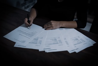 A person is signing documents on a dark wooden table. Multiple sheets of paper are spread out, with text and tables visible on them. The person is holding a black pen and wearing dark clothing, creating a contrast with the white papers.