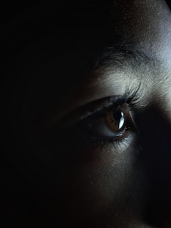 Close-up of a human eye with prominent eyelashes, surrounded by a dimly lit area. The light highlights the texture of the skin and the detail of the iris.