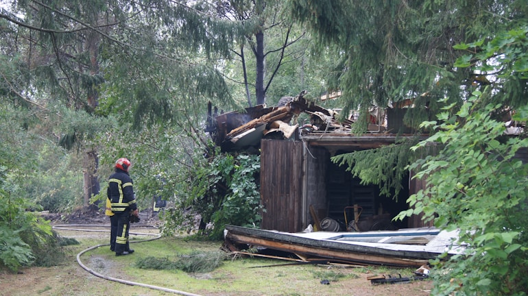 A firefighter is observing a wooden structure that has partially collapsed, surrounded by trees and greenery. The building appears damaged, with debris scattered around, likely from a recent incident. The firefighter is wearing protective gear, including a helmet and reflective clothing.
