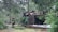 A firefighter is observing a wooden structure that has partially collapsed, surrounded by trees and greenery. The building appears damaged, with debris scattered around, likely from a recent incident. The firefighter is wearing protective gear, including a helmet and reflective clothing.