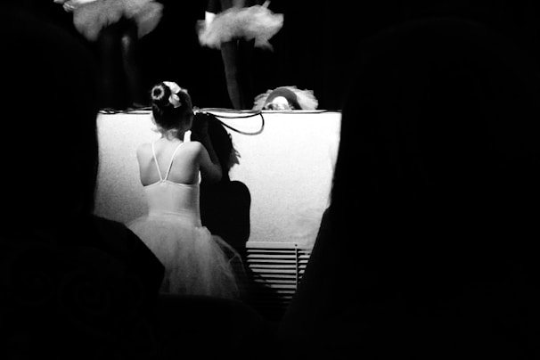 A child in a tutu is kneeling at the edge of a stage, watching performers in tutus above. The scene is in black and white, with a focus on the child's back and the stage. Shadows create a dramatic effect.