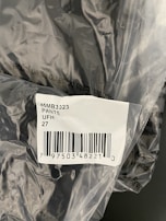 The image displays a plastic packaging containing a product labelled with a barcode sticker. The label includes text and numbers, specifically indicating '45MB3023 PANTS UFH 27' along with a barcode and a number below it.