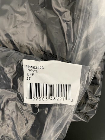 The image displays a plastic packaging containing a product labelled with a barcode sticker. The label includes text and numbers, specifically indicating '45MB3023 PANTS UFH 27' along with a barcode and a number below it.