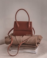 A stylish brown handbag with a structured design and looped handle, placed on top of neatly folded beige and white clothing on a neutral-colored surface.