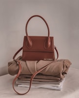 A stylish brown handbag with a structured design and looped handle, placed on top of neatly folded beige and white clothing on a neutral-colored surface.