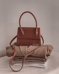A stylish brown handbag with a structured design and looped handle, placed on top of neatly folded beige and white clothing on a neutral-colored surface.