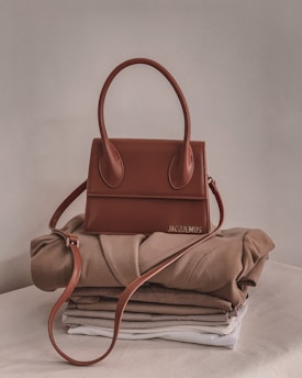 A stylish brown handbag with a structured design and looped handle, placed on top of neatly folded beige and white clothing on a neutral-colored surface.