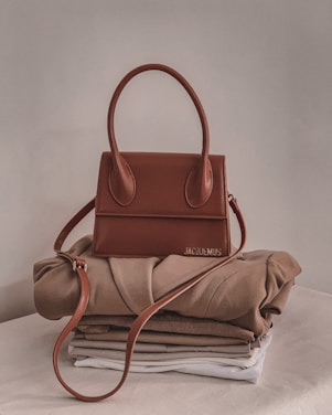 A stylish brown handbag with a structured design and looped handle, placed on top of neatly folded beige and white clothing on a neutral-colored surface.
