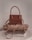 A stylish brown handbag with a structured design and looped handle, placed on top of neatly folded beige and white clothing on a neutral-colored surface.