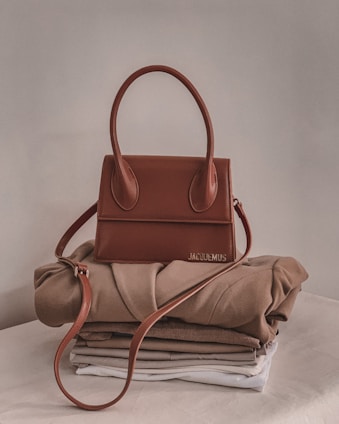 A stylish brown handbag with a structured design and looped handle, placed on top of neatly folded beige and white clothing on a neutral-colored surface.