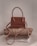 A stylish brown handbag with a structured design and looped handle, placed on top of neatly folded beige and white clothing on a neutral-colored surface.