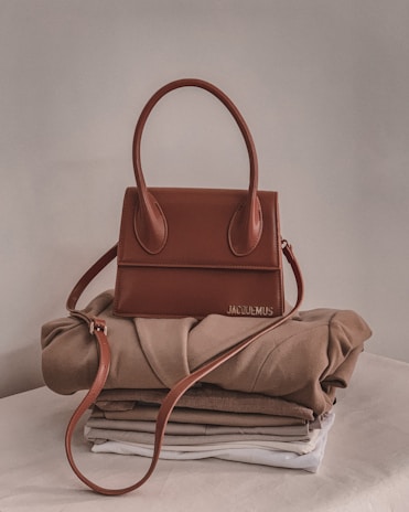 A stylish brown handbag with a structured design and looped handle, placed on top of neatly folded beige and white clothing on a neutral-colored surface.