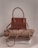 A stylish brown handbag with a structured design and looped handle, placed on top of neatly folded beige and white clothing on a neutral-colored surface.