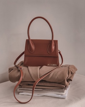 A stylish brown handbag with a structured design and looped handle, placed on top of neatly folded beige and white clothing on a neutral-colored surface.