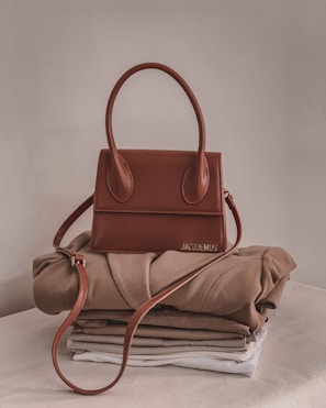 A stylish brown handbag with a structured design and looped handle, placed on top of neatly folded beige and white clothing on a neutral-colored surface.