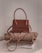 A stylish brown handbag with a structured design and looped handle, placed on top of neatly folded beige and white clothing on a neutral-colored surface.
