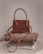 A stylish brown handbag with a structured design and looped handle, placed on top of neatly folded beige and white clothing on a neutral-colored surface.