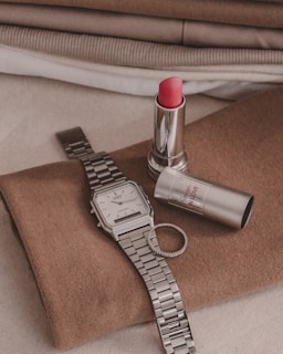 A wristwatch with a silver metal band, a ring, and a tube of red lipstick are placed on a brown piece of fabric. The fabric is layered with other textiles in the background, which are in neutral tones of beige, brown, and white.