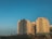 A series of modern, multi-story residential buildings with curved balconies stand under a clear blue sky. The buildings are surrounded by palm trees and are lit by the warm, golden light of the setting sun, casting distinct shadows.