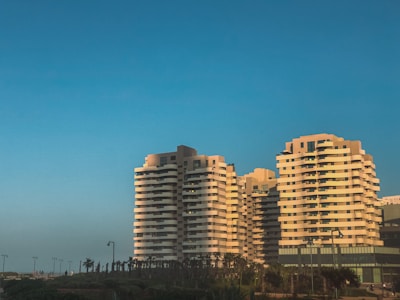 A series of modern, multi-story residential buildings with curved balconies stand under a clear blue sky. The buildings are surrounded by palm trees and are lit by the warm, golden light of the setting sun, casting distinct shadows.