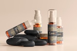 A collection of cosmetic bottles and containers are arranged artfully alongside smooth black stones. The containers are sleek, with elegant labels displaying ornate designs in golden hues against a dark background. The arrangement and lighting create a serene and luxurious aesthetic.