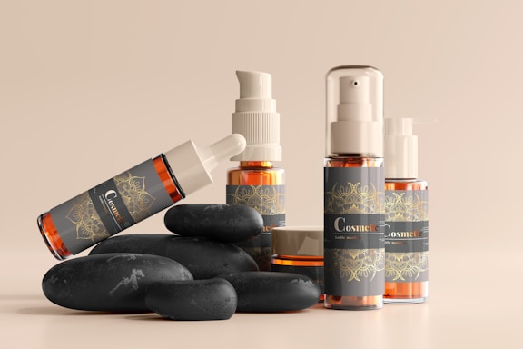 A collection of cosmetic bottles and containers are arranged artfully alongside smooth black stones. The containers are sleek, with elegant labels displaying ornate designs in golden hues against a dark background. The arrangement and lighting create a serene and luxurious aesthetic.