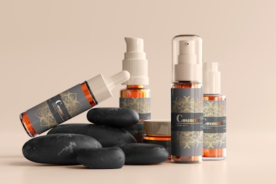 A collection of cosmetic bottles and containers are arranged artfully alongside smooth black stones. The containers are sleek, with elegant labels displaying ornate designs in golden hues against a dark background. The arrangement and lighting create a serene and luxurious aesthetic.