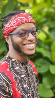A person is smiling while wearing a vibrant red bandana and bold-patterned clothing. The background is filled with lush green foliage, creating a lively and fresh atmosphere.