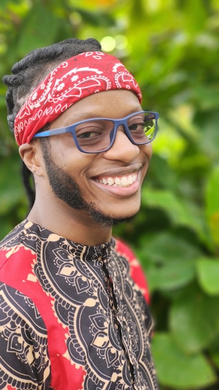 A person is smiling while wearing a vibrant red bandana and bold-patterned clothing. The background is filled with lush green foliage, creating a lively and fresh atmosphere.