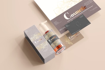 A coordinated set of cosmetic product branding materials includes a pump bottle, a rectangular box, business cards, and a leaflet. The design features elegant floral patterns in gold over grey and beige backgrounds, with the word 