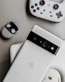 A white smartphone with a black camera bar lies on a textured surface next to wireless earbuds in a white case and a white gaming controller. The scene includes a close-up of a wooden disc with 'AUX' and 'STAND' inscribed.