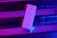 A three-button remote control with labels for ON, OFF, and a brightness icon is positioned against a background illuminated by vibrant pink and blue lighting. The remote is standing against a metallic or reflective surface with shadows and light reflections creating a dynamic visual effect.