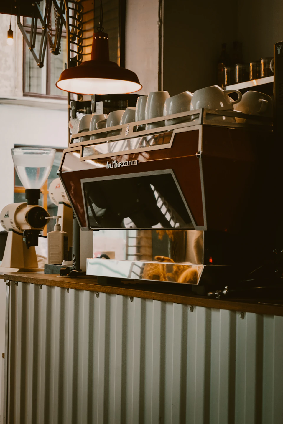 The image shows a red and white espresso machine with a black top, standing on a wooden countertop. The espresso machine is surrounded by an array of coffee cups and mugs neatly arranged on the countertop. In the background, there are shelves filled with various items, suggesting that this scene takes place in a commercial setting such as a café or restaurant.