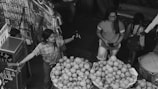 A market scene with multiple people engaged in various activities. A woman in a plaid shirt appears to be using a two-way radio, while another woman in shorts and glasses holds a smartphone and stands near a stack of fruit, predominantly pomegranates.