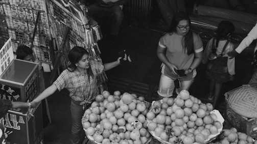 A market scene with multiple people engaged in various activities. A woman in a plaid shirt appears to be using a two-way radio, while another woman in shorts and glasses holds a smartphone and stands near a stack of fruit, predominantly pomegranates.