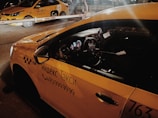 Two yellow taxis are parked under a barrier in a dimly lit area, with signage in Cyrillic script indicating they are Yandex taxis. The interior of the nearest taxi is partially visible through the side window.