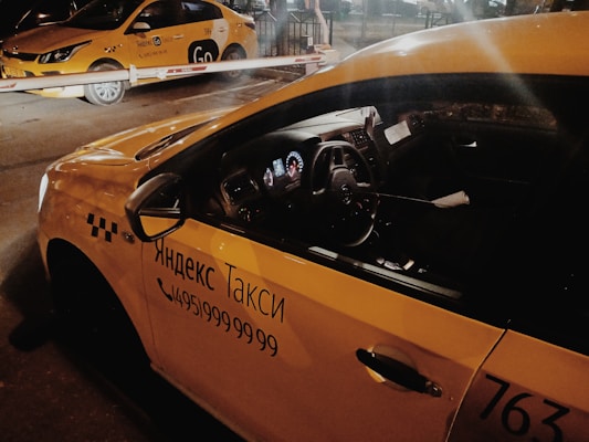 Two yellow taxis are parked under a barrier in a dimly lit area, with signage in Cyrillic script indicating they are Yandex taxis. The interior of the nearest taxi is partially visible through the side window.