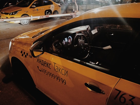 Two yellow taxis are parked under a barrier in a dimly lit area, with signage in Cyrillic script indicating they are Yandex taxis. The interior of the nearest taxi is partially visible through the side window.
