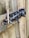 A weathered wooden gate secured with a metal slide bolt and a heavy-duty padlock. The bolt is attached to an aged, slightly rusted metal plate, and a piece of wood appears to be snapped near the latch.