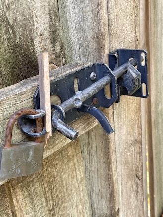 A weathered wooden gate secured with a metal slide bolt and a heavy-duty padlock. The bolt is attached to an aged, slightly rusted metal plate, and a piece of wood appears to be snapped near the latch.