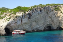A red tour boat filled with passengers floats near a rocky shoreline with large, rugged cliffs and dark caves. The clear blue water contrasts with the green shrubs growing on the cliffs.