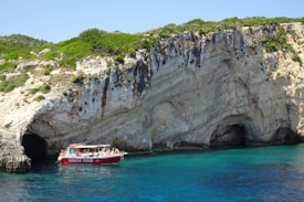 A red tour boat filled with passengers floats near a rocky shoreline with large, rugged cliffs and dark caves. The clear blue water contrasts with the green shrubs growing on the cliffs.