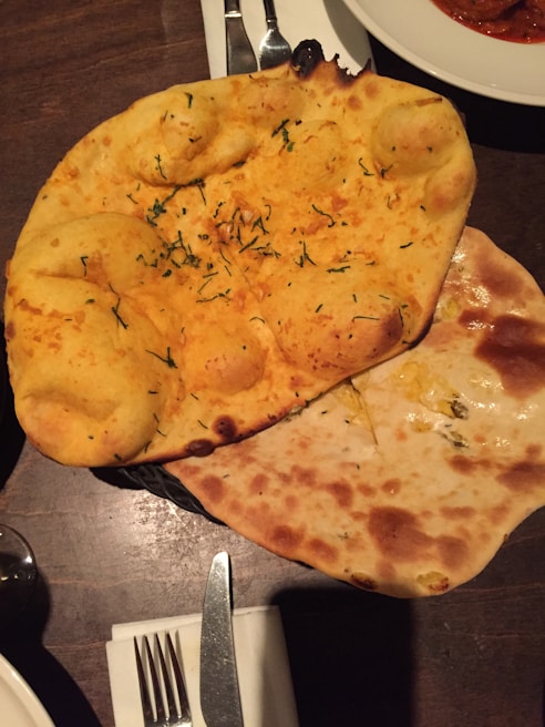 A basket of two types of naan, one topped with herbs, placed on a wooden table. Nearby, there is a fork and knife on a napkin, and a partially visible dish containing a red curry.