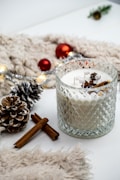 A glass candle holder containing a white candle is adorned with star anise and cinnamon sticks. The scene features a frosted pine cone, red Christmas ornaments, a green pine branch, and string lights, all placed on a white and beige fuzzy surface.