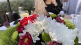 A bouquet of flowers featuring white, red, and green blooms with a wooden tree decoration in the center. In the background, there are blurred figures of people seated at a table set for an event, with glasses and foliage visible.