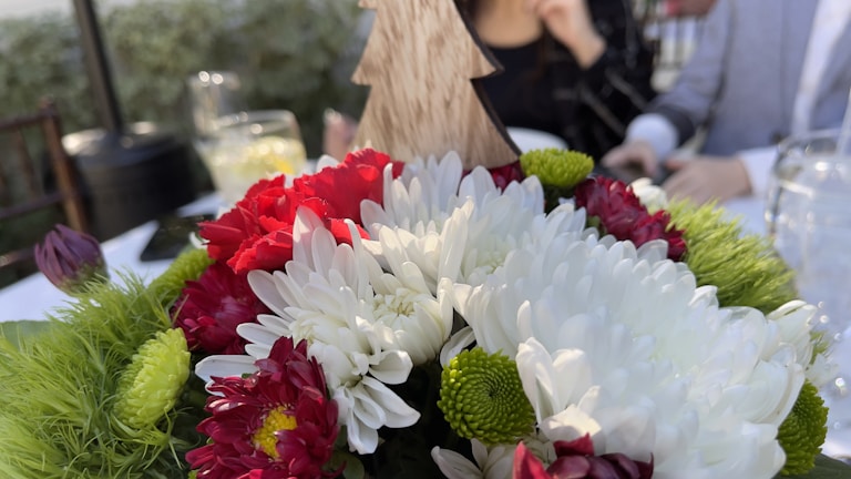 A bouquet of flowers featuring white, red, and green blooms with a wooden tree decoration in the center. In the background, there are blurred figures of people seated at a table set for an event, with glasses and foliage visible.