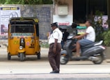 A traffic policeman stands on the side of a road near a parked yellow auto-rickshaw. A person on a scooter is passing by the policeman. The background includes a brick wall with posters, some greenery, and a partially visible commercial establishment.