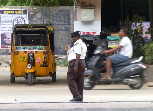 A traffic policeman stands on the side of a road near a parked yellow auto-rickshaw. A person on a scooter is passing by the policeman. The background includes a brick wall with posters, some greenery, and a partially visible commercial establishment.