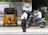 A traffic policeman stands on the side of a road near a parked yellow auto-rickshaw. A person on a scooter is passing by the policeman. The background includes a brick wall with posters, some greenery, and a partially visible commercial establishment.